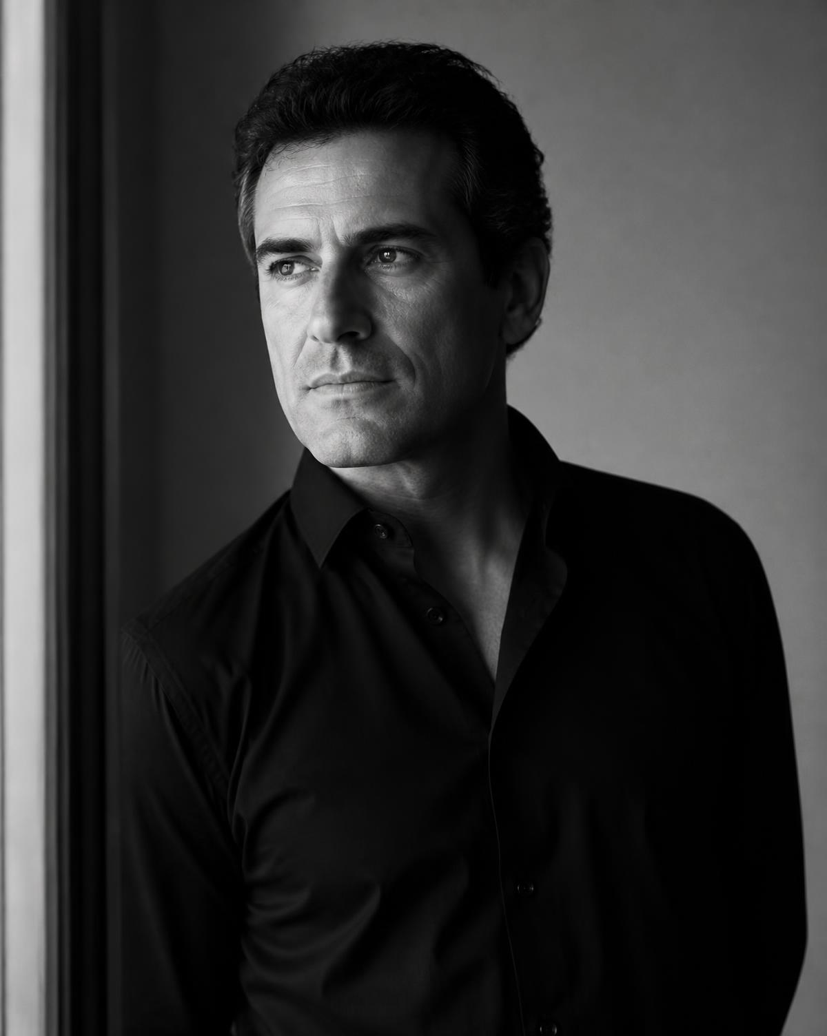 Editorial black and white portrait of Manuel Serrano Ortega beside a window